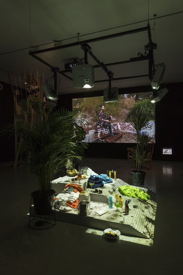 Installation view of TARO IZUMI’s Spring Is Between Winter and Summer, Sleeping Is Between Spring and Early Summer, 2018, concrete stage with plants, fruit, drinks, stained glass sunglasses and beach apparel, looped video projection and video on monitor, dimensions variable, at “My Eyes Are Not in the Centre,” White Rainbow, London, 2018.